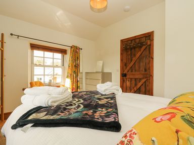 A bedroom with a bed folded towels a wooden door a dresser a window with floral curtains and a yellow chair at Lower Gadles Burnthouse near Falmouth