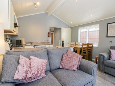An open living room and kitchen area with gray sofas with pink cushions a wooden dining table with chairs and kitchen appliances at Wansfell Lodge Pony Meadow 36 White Cross Bay near Windermere