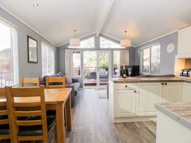 A dining table with four chairs a sofa kitchen island with a bottle and glass large windows and glass door leading to a deck at Wansfell Lodge Pony Meadow 36 White Cross Bay near Windermere