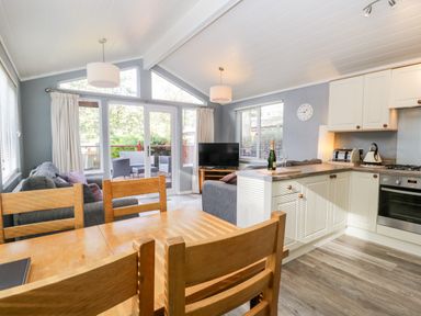 A dining table with wooden chairs next to a kitchen area and living room with sofas and a television at Wansfell Lodge Pony Meadow 36 White Cross Bay near Windermere