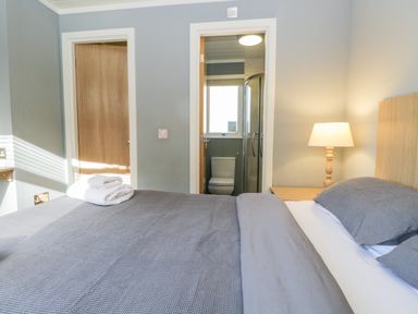 A bedroom with a bed covered with gray bedding a bedside table with a lamp two doors one leading to a bathroom at Wansfell Lodge Pony Meadow 36 White Cross Bay near Windermere