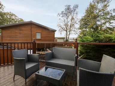 A wooden deck with outdoor seating including chairs and a table and a wooden cabin in the background at Wansfell Lodge Pony Meadow 36 White Cross Bay near Windermere