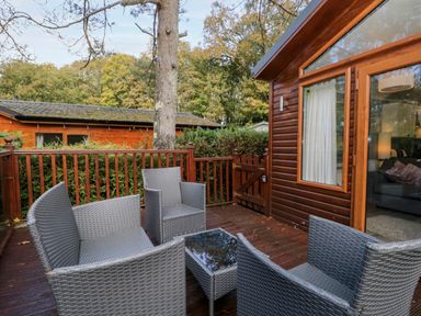 An outdoor wooden deck with grey wicker chairs and a small table next to a wooden house at Wansfell Lodge Pony Meadow 36 White Cross Bay near Windermere