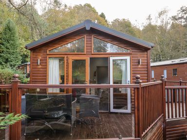 A wooden cabin with glass doors and a deck with outdoor chairs surrounded by trees at Wansfell Lodge Pony Meadow 36 White Cross Bay near Windermere