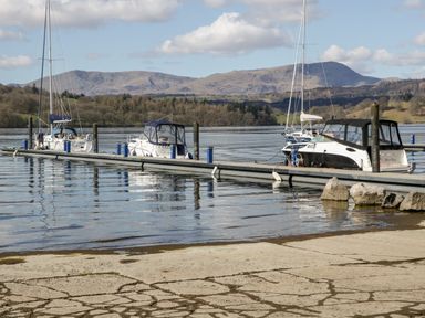 A dock with several boats moored on a lake with hills and trees in the background at Wansfell Lodge in Pony Meadow 36 White Cross Bay near Windermere