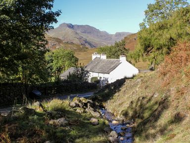 A white house near a road with trees and a small stream in a mountainous landscape at Wansfell Lodge in Pony Meadow 36 White Cross Bay near Windermere