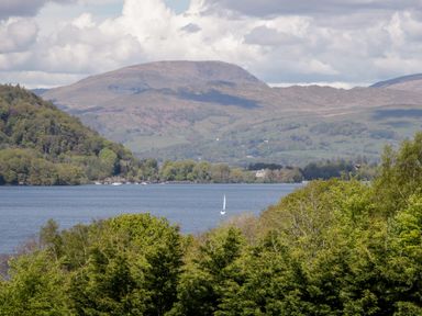 A lake with a sailboat surrounded by hills and trees at Wansfell Lodge Pony Meadow 36 White Cross Bay near Windermere