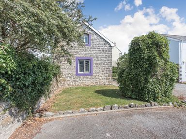 A stone house with purple window frames surrounded by bushes and a lawn at The Coach House in St Florence