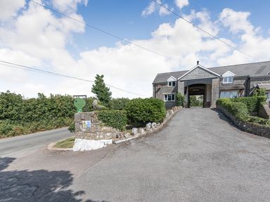 A driveway leading to a stone building with an archway surrounded by hedges at The Coach House in St Florence