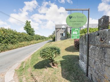 A road beside a grassy area with a sign for Ivy Tower Village Private Holiday Cottages and electric vehicle charging with a stone wall and building in the background at The Coach House in St Florence