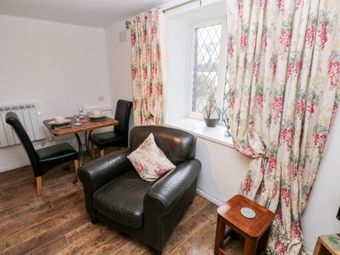 A small dining area with a wooden table set for two and a black leather armchair near a window with floral curtains at The Coach House in St Florence
