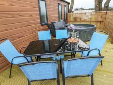 An outdoor patio with a glass table surrounded by six blue chairs and a hot tub next to a wooden wall and fence at Oyster Lodge in Broad Haven