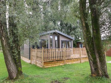 A wooden cabin with a fenced porch surrounded by trees and grass at Coniston View 9 Dock Acres near Tewitfield