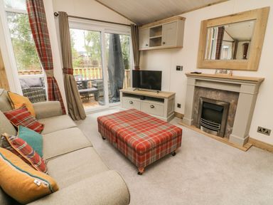 A living room with a sofa with cushions a plaid ottoman a fireplace a television on a cabinet and sliding glass doors at Coniston View 9 Dock Acres near Tewitfield