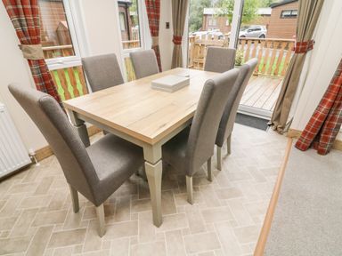 A dining area with a rectangular wooden table and six upholstered chairs near windows with plaid curtains at Coniston View 9 Dock Acres near Tewitfield