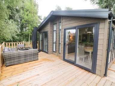 An outdoor wooden deck with wicker seating and glass table attached to a house with large sliding glass doors at Coniston View 9 Dock Acres near Tewitfield