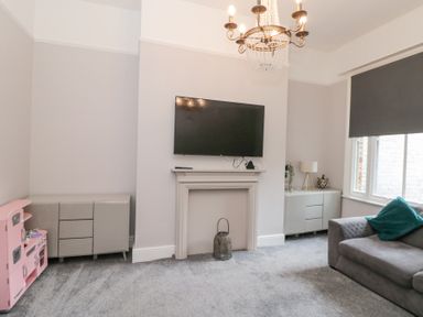 A living room with a wall mounted TV above a fireplace two cabinets a gray sofa and a pink toy kitchen at Westbourne House in Scarborough