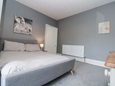 A bedroom with a gray bed frame, a bedside table with a lamp, gray walls, two pieces of wall art, a radiator, and a wooden dresser at Westbourne House in Scarborough