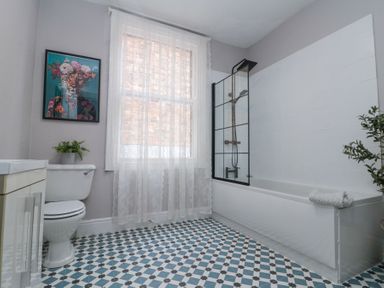 A bathroom with a bathtub and shower screen a toilet with a plant on top a window with lace curtains and patterned tiled floor at Westbourne House in Scarborough