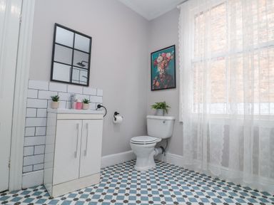 A bathroom with a toilet, a sink cabinet with two small plants, a mirror, patterned floor tiles, a window with lace curtains, and a framed picture of a tiger at Westbourne House in Scarborough