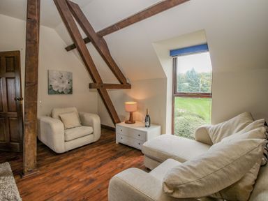 A sitting area with a beige armchair and sofa near a window with a wooden floor and exposed wooden beams at Owlbury Hall Barn in Snead near Bishops Castle Montgomery
