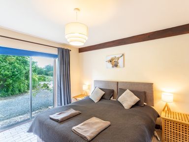 A bedroom with a double bed, two pillows, two folded towels, bedside tables with lamps, and a sliding glass door showing greenery outside at Owlbury Hall Barn in Snead near Bishop's Castle Montgomery