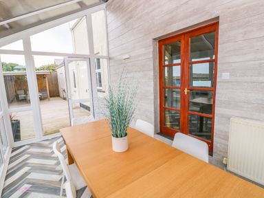 A dining table with white chairs and a plant in a glass conservatory with wooden doors at Ocean House in Broad Haven
