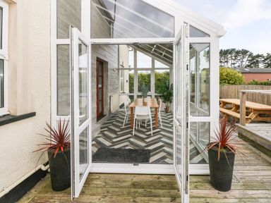 A dining area with a wooden table and four white chairs inside a glass enclosed porch at Ocean House in Broad Haven