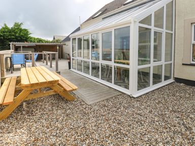An outdoor area with a wooden picnic table a glass conservatory and a deck with blue chairs at Ocean House in Broad Haven