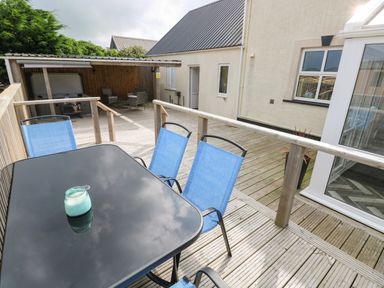 An outdoor wooden deck with a glass table and blue chairs a covered seating area and a building with windows and a door at Ocean House in Broad Haven