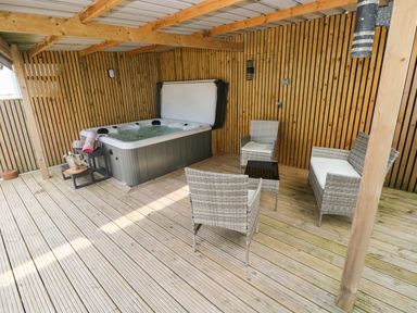 A covered wooden deck with a hot tub sofa chairs and a small table at Ocean House in Broad Haven