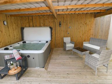 A covered outdoor area with a hot tub towels a small table with a wine bottle and glasses and wicker seating at Ocean House in Broad Haven