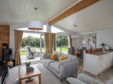 A living room with a grey sofa and yellow cushions next to sliding glass doors leading to a patio with outdoor seating at Fairview Lodge in Pwllheli