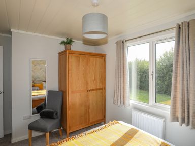 A bedroom with a wooden wardrobe a gray chair a tall mirror and a window with curtains at Fairview Lodge in Pwllheli