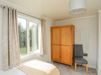 A bedroom with a window and curtains a wooden wardrobe a gray chair and a full length mirror at Fairview Lodge in Pwllheli