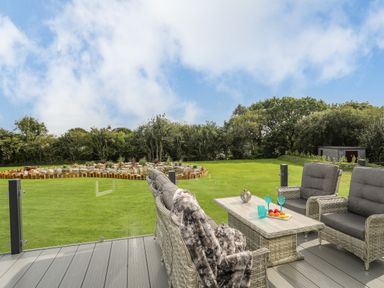 An outdoor patio with wicker chairs and a table overlooking a lawn and garden at Fairview Lodge in Pwllheli