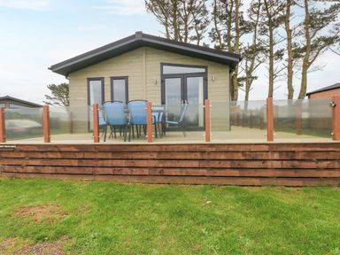 A wooden lodge with glass patio doors and a deck with blue chairs and a table at Driftwood Lodge in Hasguard Cross near Broad Haven