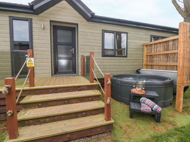 A wooden deck with steps leading to a door of a building next to a hot tub with towels and glasses on a small black stand at Driftwood Lodge Hasguard Cross near Broad Haven