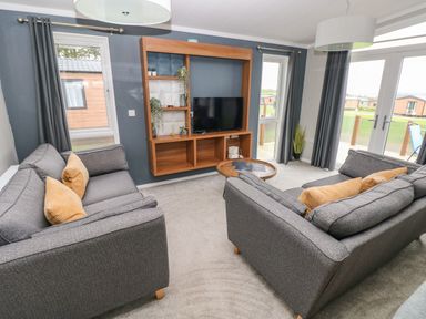 A living room with two grey sofas with yellow cushions a wall-mounted wooden shelving unit with a TV a round wooden coffee table and large windows at Driftwood Lodge in Hasguard Cross near Broad Haven