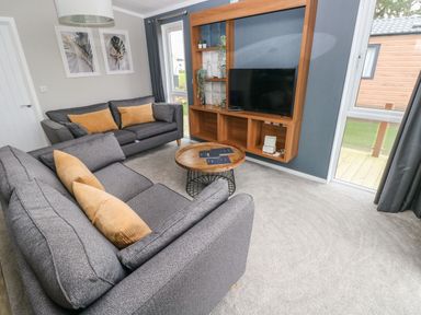 A living room with grey sofas and brown cushions a round wooden coffee table and a wall-mounted wooden entertainment unit with a TV at Driftwood Lodge in Hasguard Cross near Broad Haven
