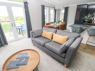 A living room with a grey sofa and yellow cushions a round wooden coffee table and a dining table with brown chairs at Driftwood Lodge in Hasguard Cross near Broad Haven