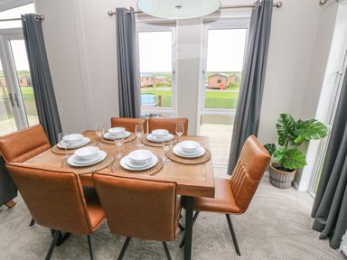 A dining area with a wooden table set for six with brown chairs and windows with gray curtains at Driftwood Lodge Hasguard Cross near Broad Haven
