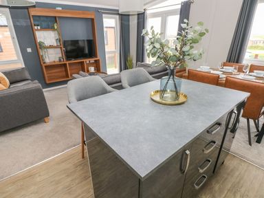 A living area with a kitchen island in the foreground a vase with branches on the island a grey sofa and armchairs a mounted wooden TV unit a dining table set with plates and glasses at Driftwood Lodge Hasguard Cross near Broad Haven