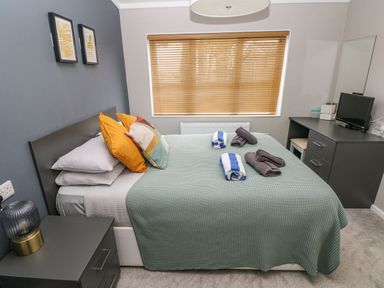 A bedroom with a bed covered with a green blanket and rolled towels two framed pictures above the bed a bedside table with a lamp and a desk with a small TV at Driftwood Lodge in Hasguard Cross near Broad Haven
