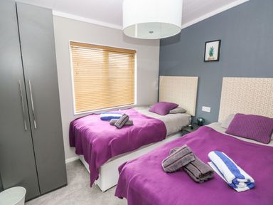 A bedroom with two single beds purple blankets pillows a wardrobe and a window with blinds at Driftwood Lodge in Hasguard Cross near Broad Haven