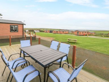 An outdoor deck with a table and six chairs overlooking a grassy area with multiple small lodges at Driftwood Lodge in Hasguard Cross near Broad Haven