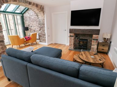 A living room with a blue sofa a round wooden coffee table a brick fireplace with a black stove two yellow chairs near a stone wall window at Skomer Cottage Sandy Haven near Milford Haven
