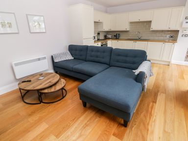 A living room with a blue sectional sofa a wooden nesting coffee table and a kitchen with white cabinets at Skomer Cottage in Sandy Haven near Milford Haven