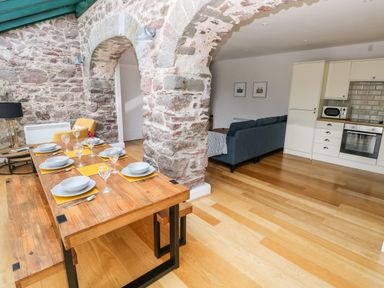 A dining table set for six in a room with stone arches and a kitchen and living area visible at Skomer Cottage in Sandy Haven near Milford Haven