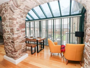 A dining area with a wooden table and benches and a sitting area with two orange chairs near stone archways and glass windows at Skomer Cottage in Sandy Haven near Milford Haven